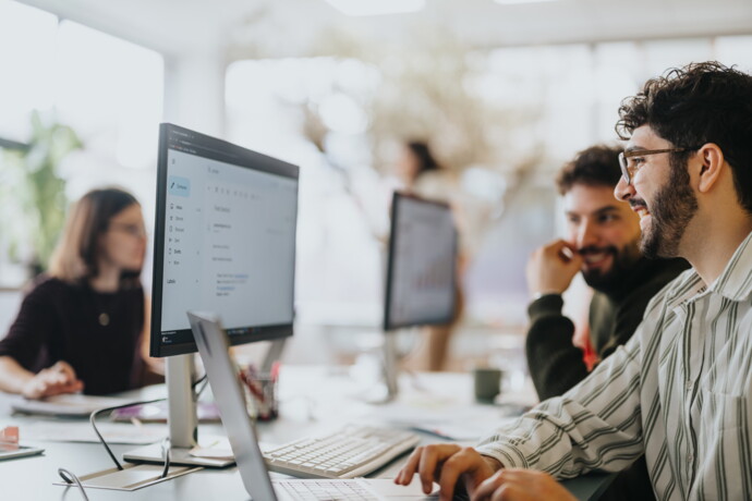 Zwei Männer lächeln und arbeiten gemeinsam an Computern, während im Hintergrund weitere Mitarbeiter an ihren Schreibtischen arbeiten.