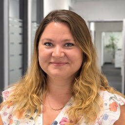 Porträtfoto einer Frau mit schulterlangen welliger Haarfarbe, die nach vorne blickt und lächelt. Sie trägt ein Oberteil mit farbigen Mustern und eine Halskette vor verschwommenem Büroinnenhintergrund