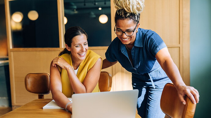 Zwei Frauen arbeiten gemeinsam an einem Laptop in einem modernen Büro. Eine sitzt in gelbem Oberteil, die andere steht daneben in dunkelblahem Hemd und Brille.